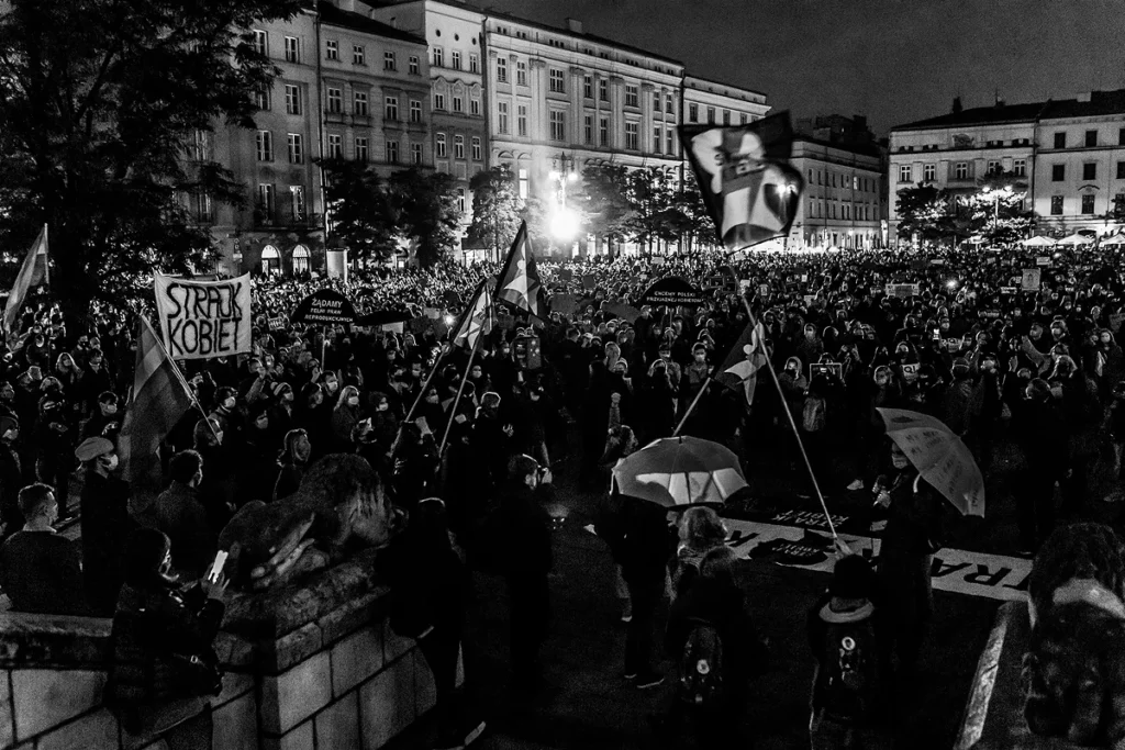 Tłum zebrany wieczorem na rynku głównym w Krakowie wymachuje flagami i transparentami Strajku Kobiet. W tle widać kamienice a ludzie zajmują cały obszar rynku.