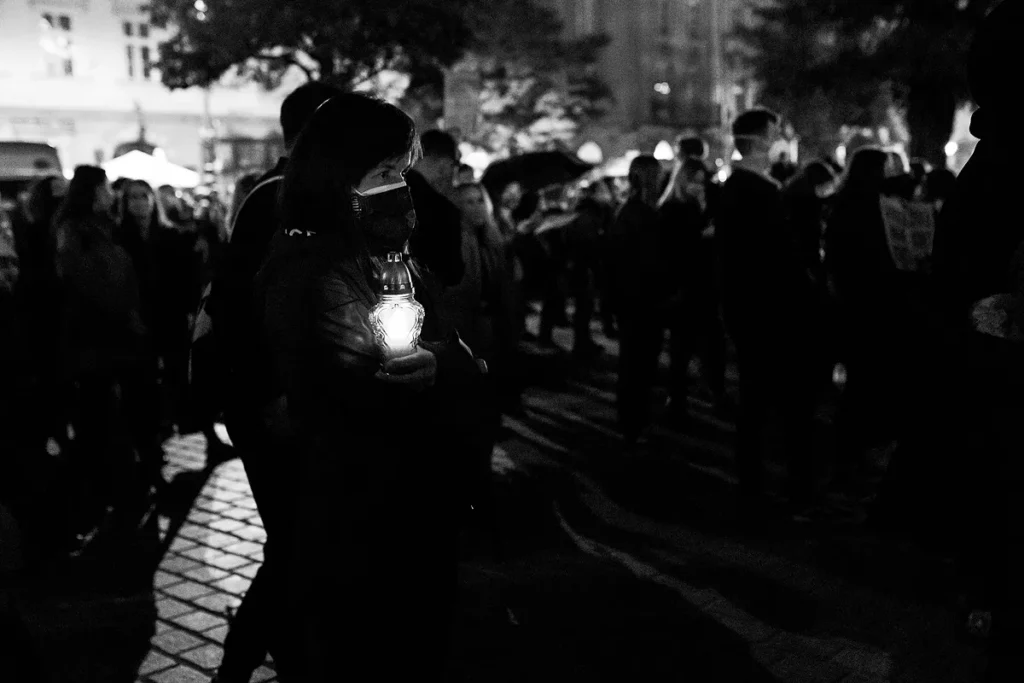 na pierwszym planie znajduje się kobieta ubrana w zimową odzież. Przytula do swojej klatki piersiowej zapalony znicz. W tle znajdują się inni ludzie. Na zdjęciu dominują czernie a znicz pełni funkcję bardzi jasnego akcentu bieli.