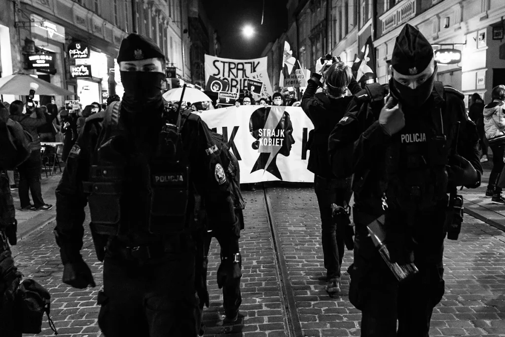 Scena przedstawia pochód demonstrantów idący wąską ulicą pomiędzy kamienicami. Na pierwszym planie widzimy postaci dwóch policjantów w mundurach bojowych, którzy idą w stronę w stronę obiektywu fotografa. Za nimi widać fragment transparentu Strajk Kobiet, który zasłania maszerujących demonstrantów. W tle flagi i transparenty z hasłami. W górnej części na środku kompozycji, na tle ciemnego nieba widoczny jest rozbłysk latarni ulicznej, który wygląda jak słońce.