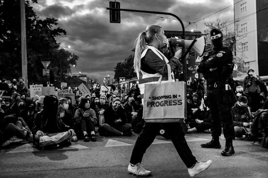 Młoda dziewczyna stoi na skrzyżowaniu dróg i przemawia do tłumu przez megafon. Na ramieniu ma torbę zakupową z napisem shopping in progress. Na drugim planie siedząca młodzież w maseczkach z powodu epidemii covid-19. Megafon jest skierowany w stronę głowy policjanta, który w bojowym umundurowaniu stoi w rozkroku i z założonymi rękami ją obserwując ją.