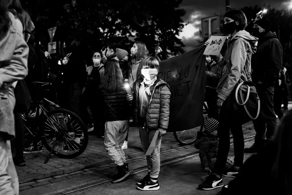 Ujęcie na demonstrantów. Na środku stoi chłopiec w wieku ok. 8 lat, obok jego mama trzyma transparent. Chłopiec spogląda w kierunku widza. Jego twarz oświetla smartfon, czyniąc jego postać najważniejszą, centralną częścią kompozycji.
