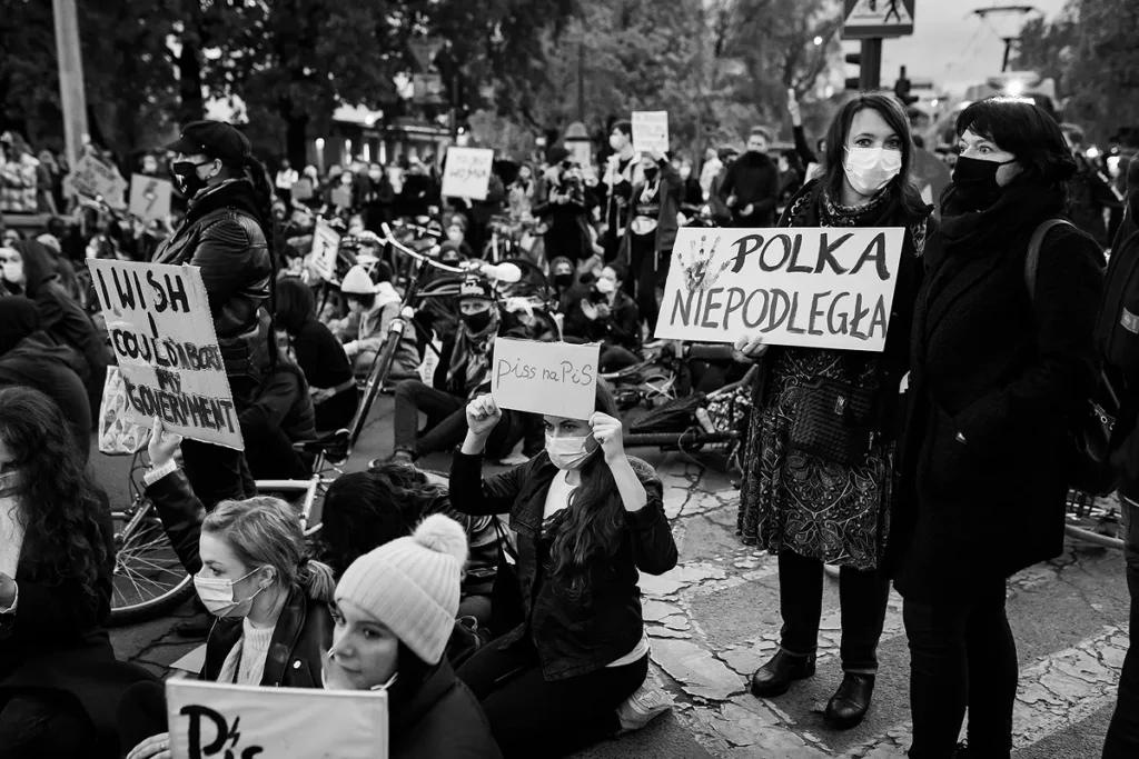 Tłum ludzi. Większość z nich siedzi na jezdni samochodowej i wznosi transparenty z hasłami przeciwko partii PIS i prawu zabraniającemu aborcji. Po prawej stronie stoją dwie kobiety. Jedna z nich spogląda w kierunku obiektywu i trzyma tabliczkę z haslem Niepodległa Polska.