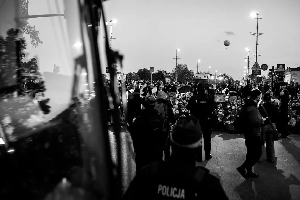 Ujęcie na tłum demonstrantów. Od lewej strony jedną trzecią część zdjęcia zajmuje tramwaj, który przez zastosowaną głębię ostrości jest nieostry. W głębi zdjęcia znajdują się siedzący na ziemi demonstranci. W dalekiej perspektywie, na tle nieba unosi się balon widokowy.