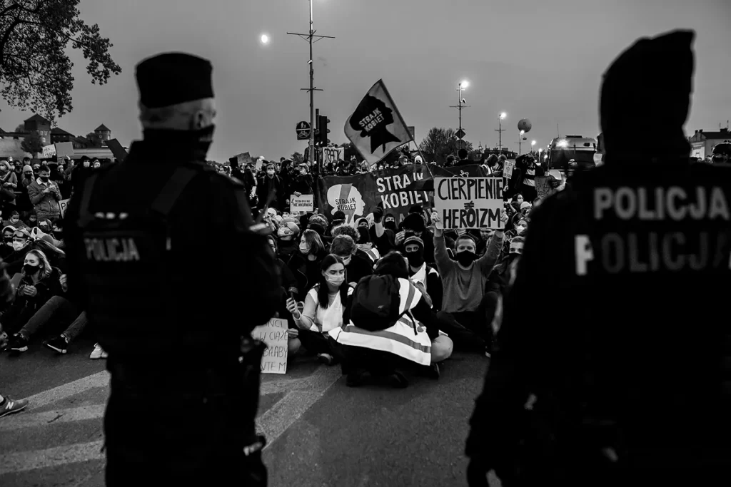 Na pierwszym planie stoi dwóch policjantów zwróconych tyłem do widza i przodem do siedzących demonstrantów, którzy trzymają transparenty z hasłami: strajk kobiet, cierpienie ≠ heroizm